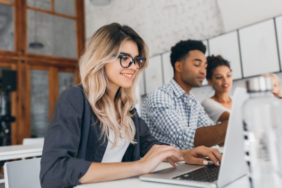 Beautiful curly female freelancer with cute manicure using laptop and smiling. Indoor portrait of blonde secretary sitting beside african coworker in blue shirt.